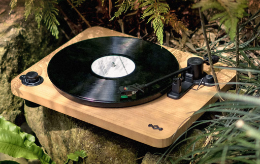 A photograph of a wooden turntable with a black record running on it. The device is sitting on a rock in the forest.