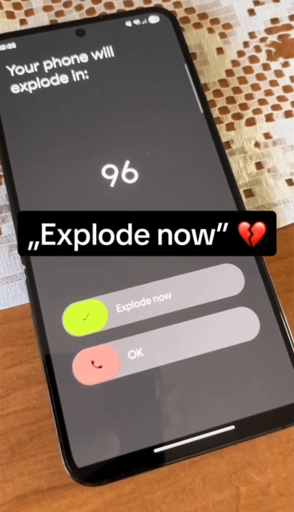 A low quality, angled photo of a Samsung phone. On the screen it says "Your phone will explode in 96 [seconds]". There are two swipe options below, Explode now, or Ok. The image has a caption with the following text: "'explode now" 💔
